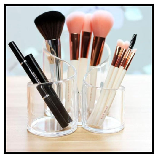 Three clear acrylic makeup brush holders on light wood surface holding makeup brushes with pink peach and black bristles