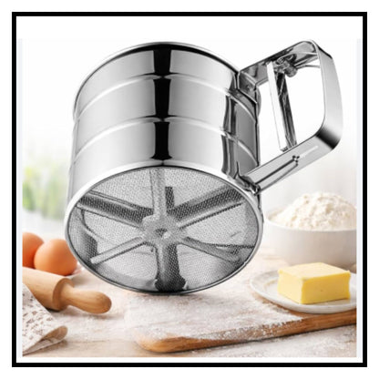Stainless steel flour sifter with handle on floured countertop with baking ingredients like flour, eggs, rolling pin and butter